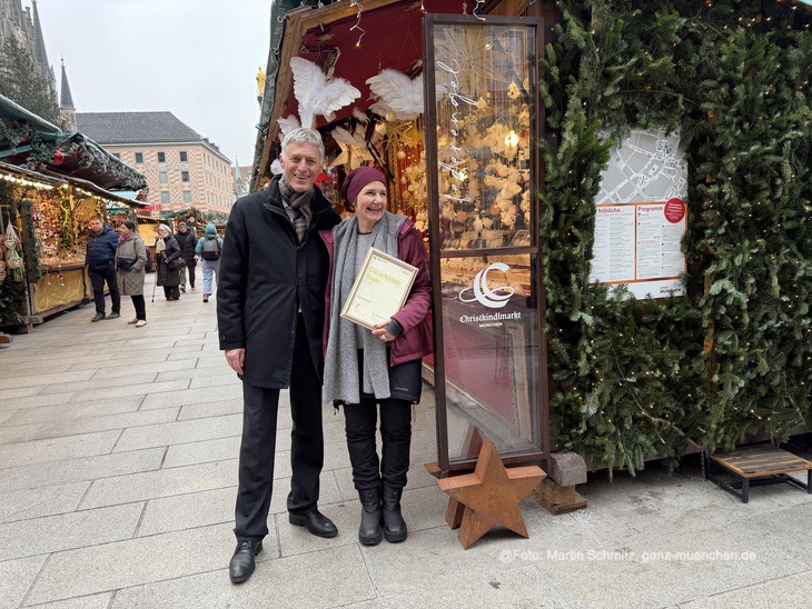 Pr&auml;miert als sch&ouml;nste Bude 2025: Michaela Trost vom Federengel auf dem M&uuml;nchner Christkindlmarkt erhielt eine Urkunde von Kurt Kapp (stellv. Leiter Referat f&uuml;r Arbeit und Wirtschaft)  (&copy;Foto: Martin Schmitz)